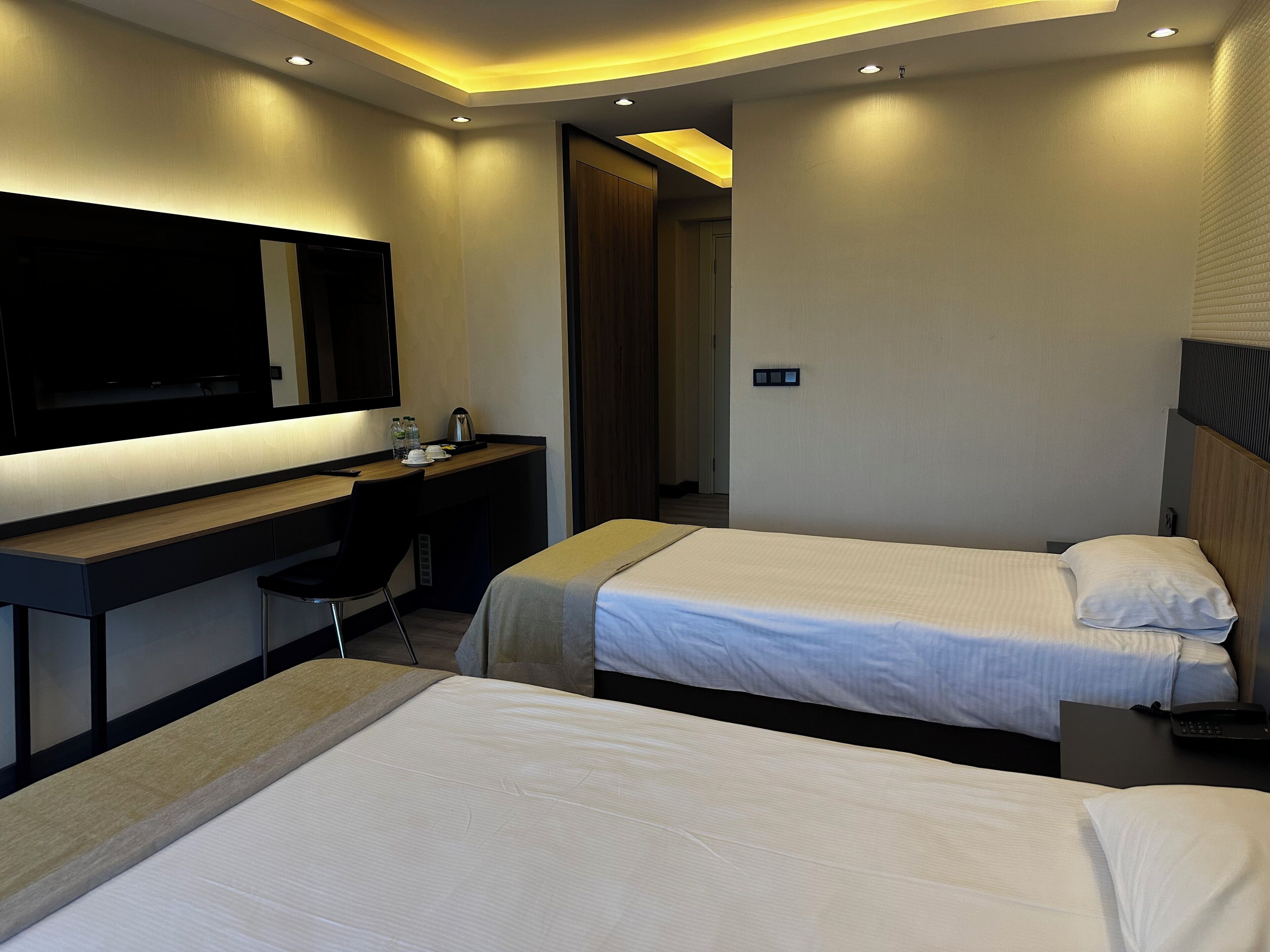 Quarto de hotel com duas camas, uma mesa com cadeira, uma TV de tela plana e uma porta que dá para outro cômodo.
