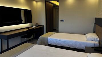 Quarto de hotel com duas camas, uma mesa com cadeira, uma TV de tela plana e uma porta que dá para outro cômodo.