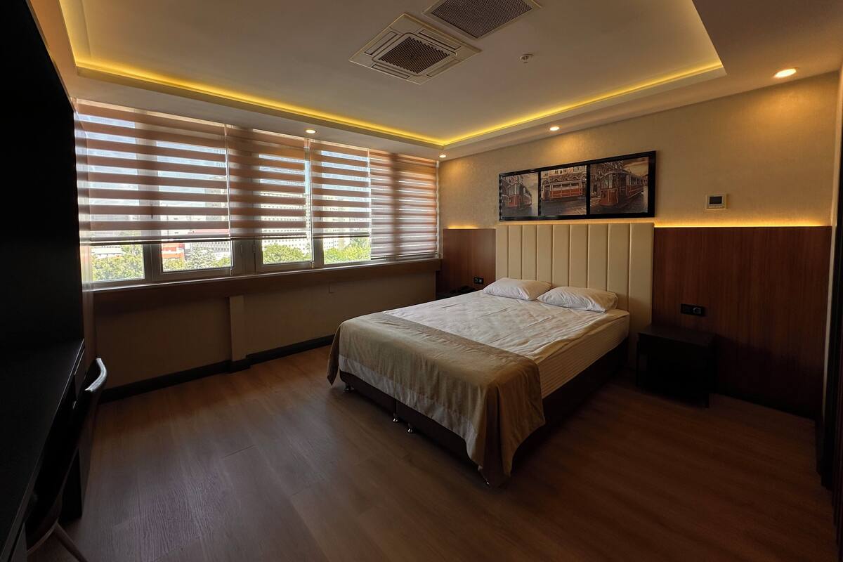 A hotel room with a bed, windows with blinds, a headboard, a wall-mounted artwork, and a ceiling vent.