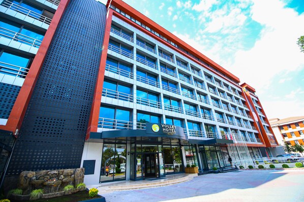 Un hotel moderno con un ingresso ben visibile, diversi piani e un cartello che riporta “Volley Hotel”.