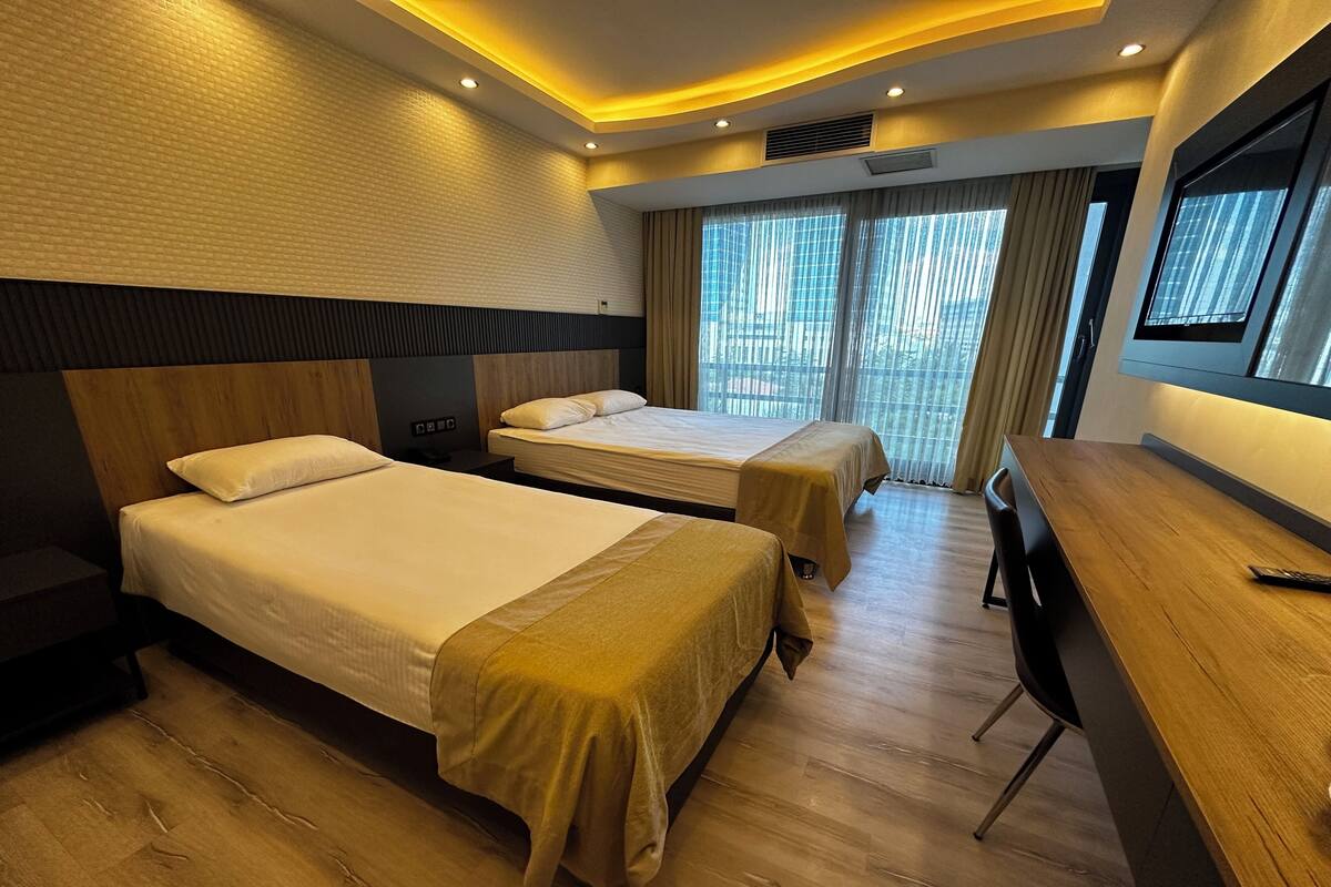 A hotel room with two beds, a wooden desk, and a large window with curtains.