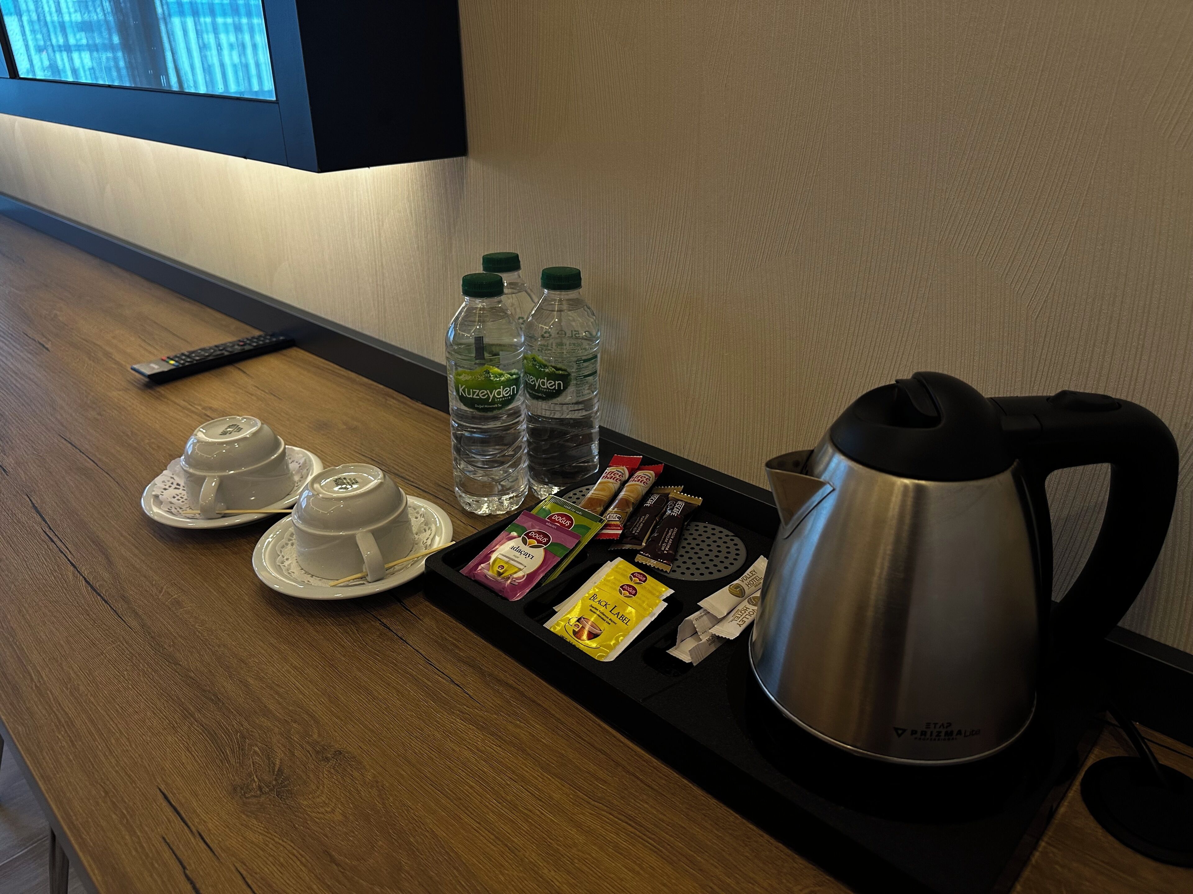 A hotel room's minibar with a tray containing a teapot, two cups, two bottles of water, a remote control, and assorted snacks.
