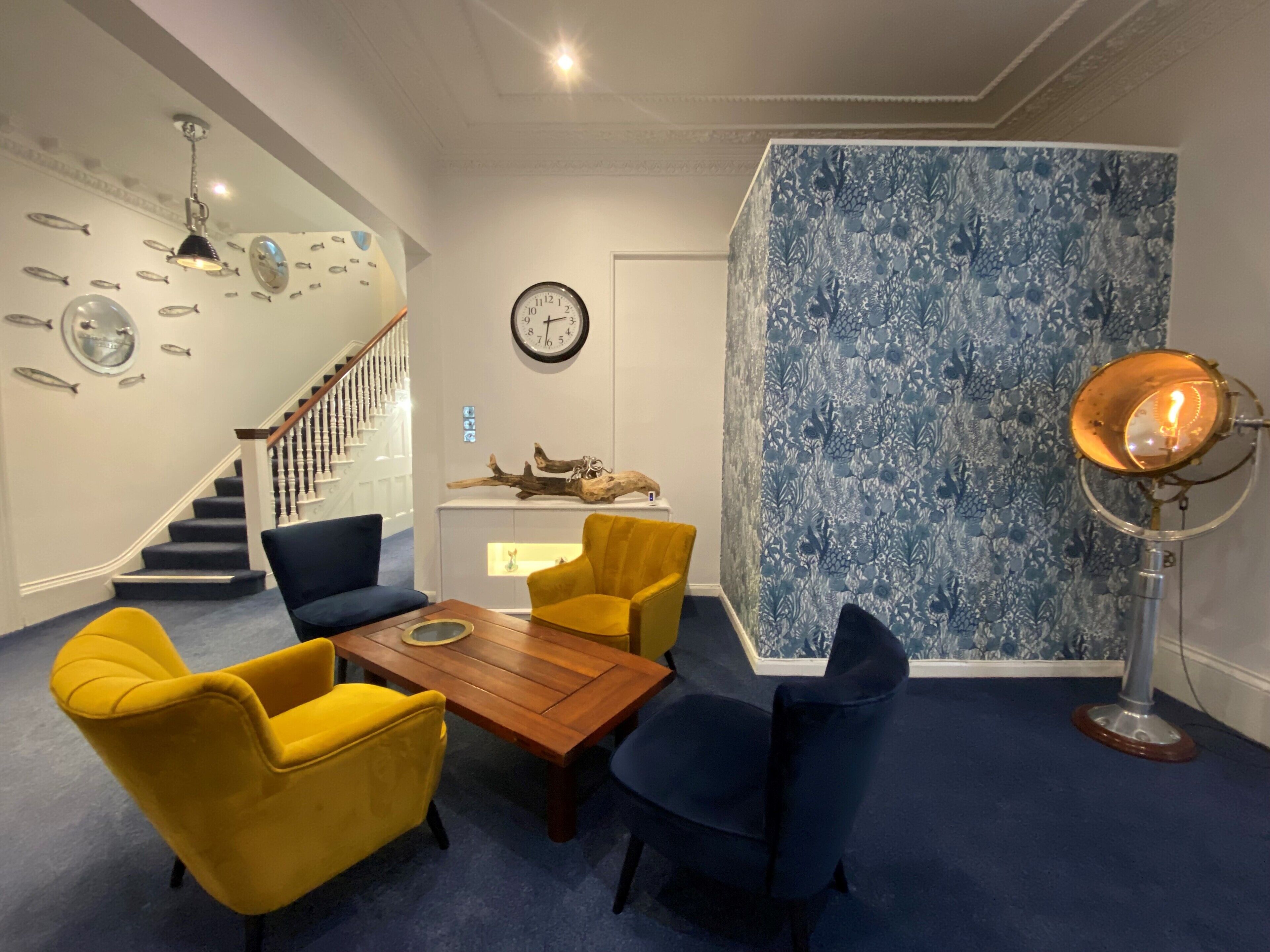 A room with a wooden coffee table, yellow and blue chairs, a staircase, a clock, and a blue patterned wall.
