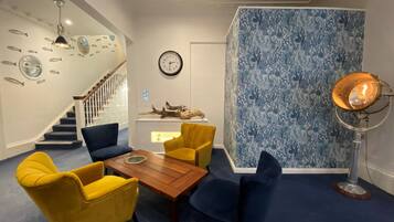 Una habitación con una mesa de centro de madera, sillas amarillas y azules, una escalera, un reloj y una pared con estampado azul.