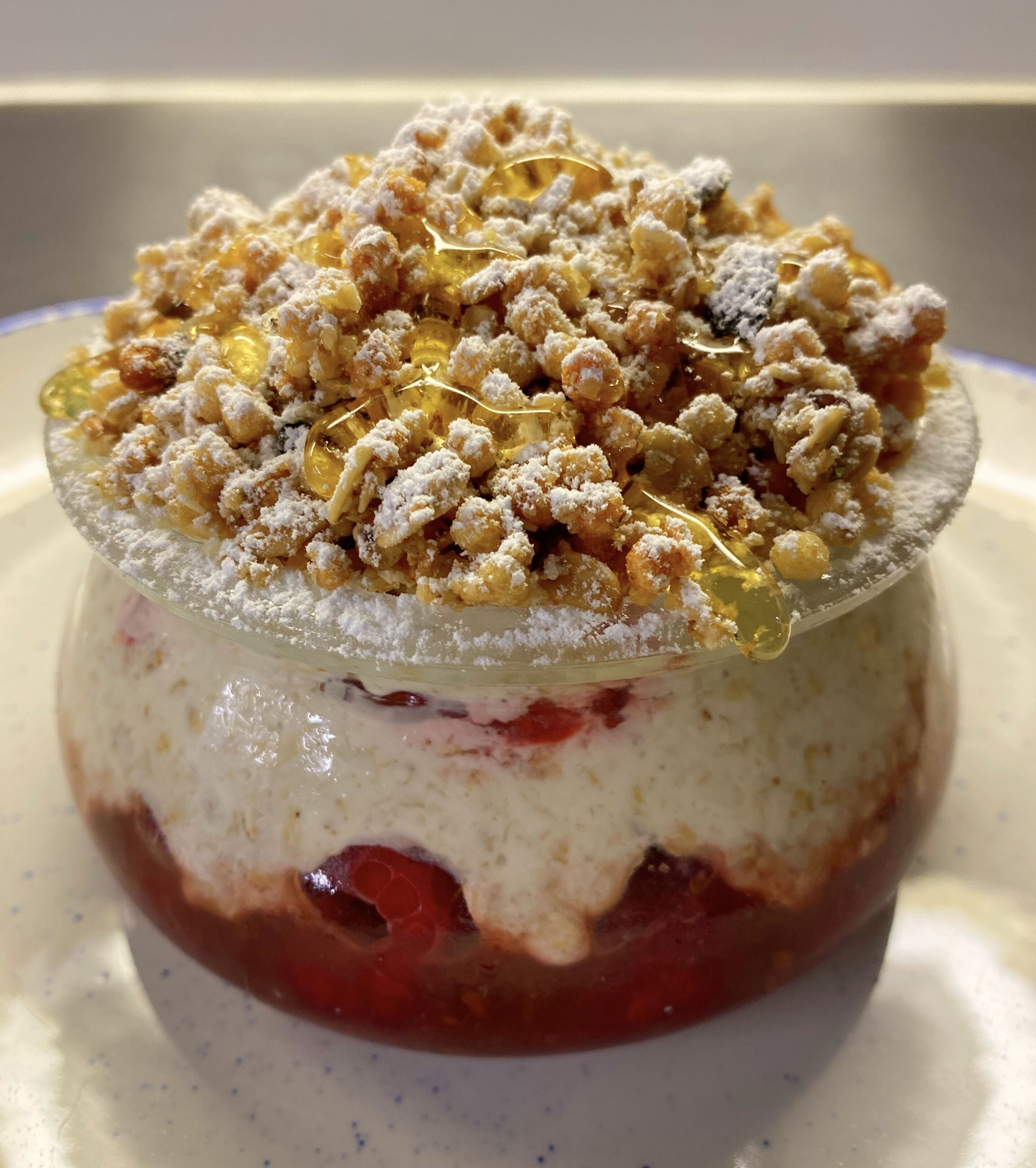 A dessert with a creamy base, a red fruit layer, and a crumbly topping, served on a clear glass dish.