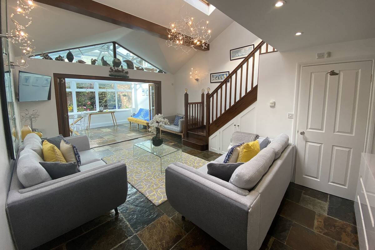 A spacious living area with a glass coffee table, two grey sofas, a wooden staircase, and a view of the outdoors.