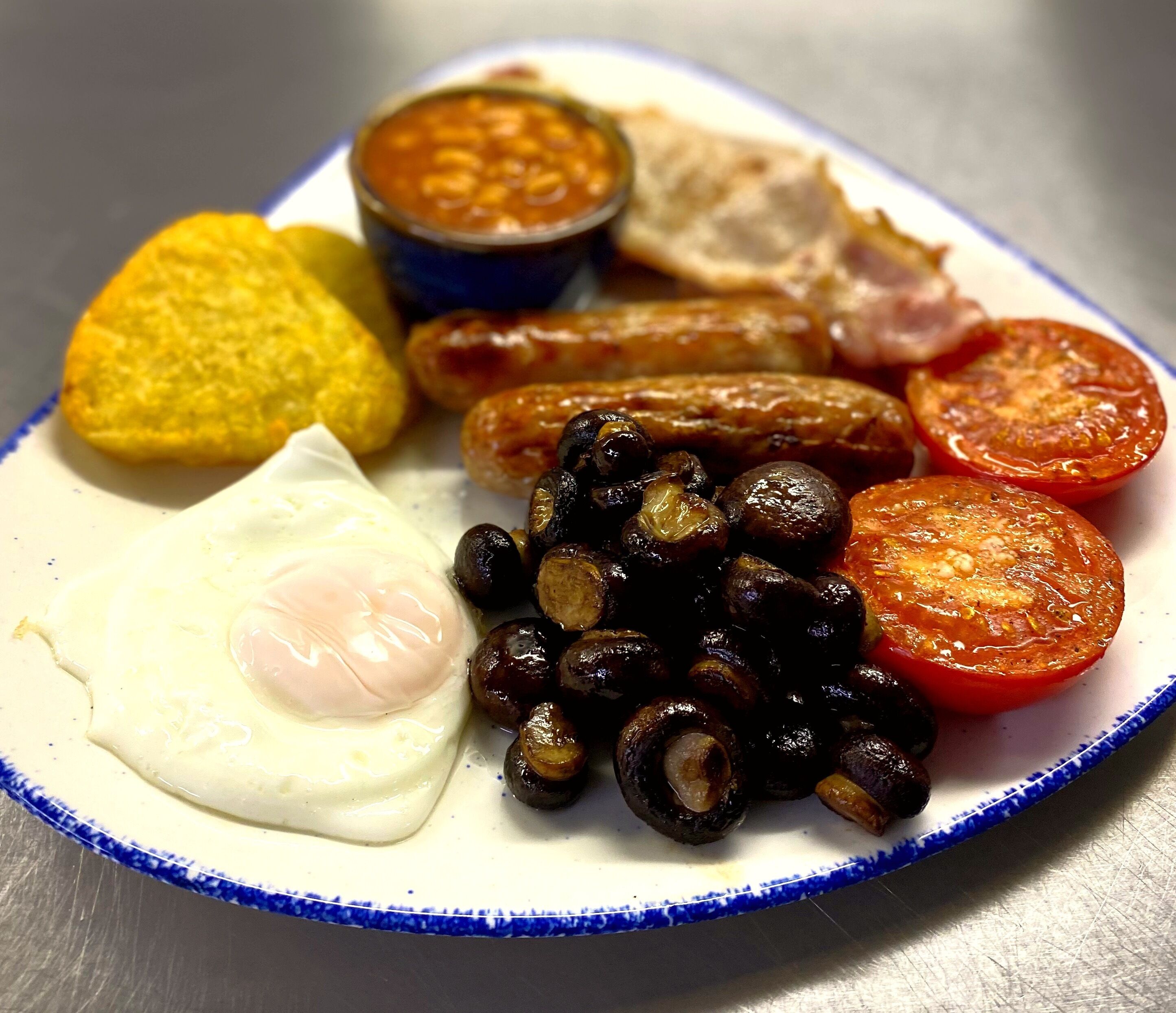 A plate with a fried egg, sausages, tomatoes, black olives, and a small bowl of beans.