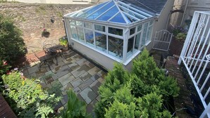 A conservatory with a glass roof, surrounded by a garden with various plants and a patio area with a table and chairs. - Rooms At Babbacombe (Torquay)
