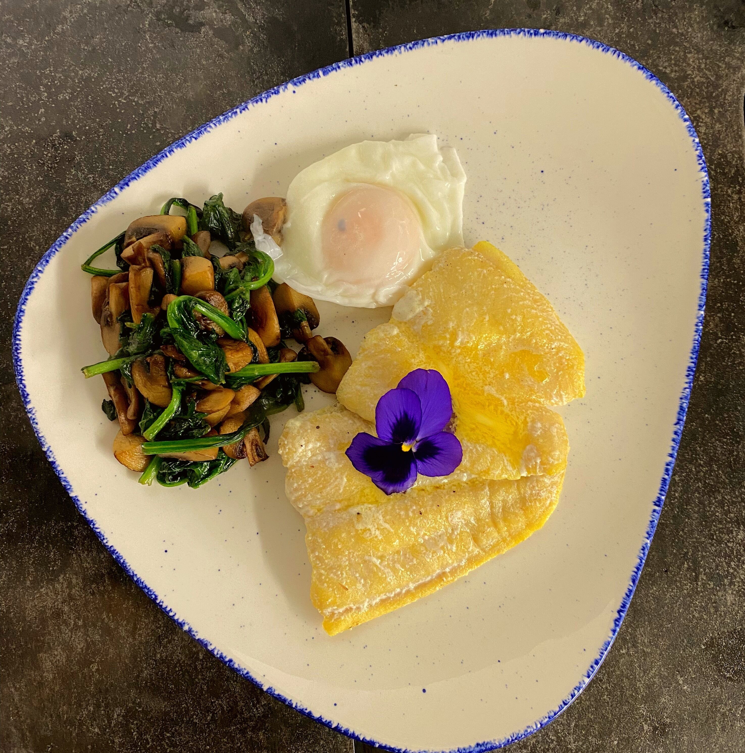 Un plato con un huevo frito, champiñones salteados y espinacas, acompañado de una rebanada de tostada dorada y una flor morada.