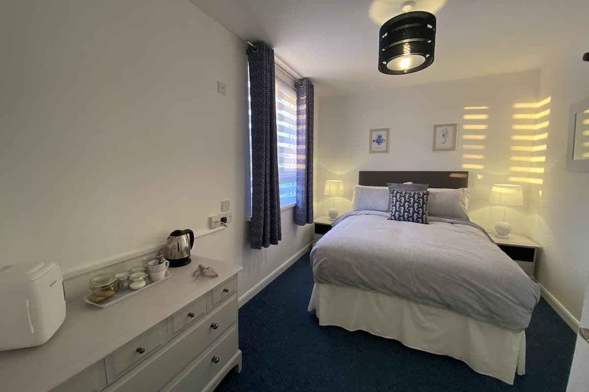 A hotel room with a bed, bedside lamps, a nightstand, a dresser, a kettle, and a window with curtains.
