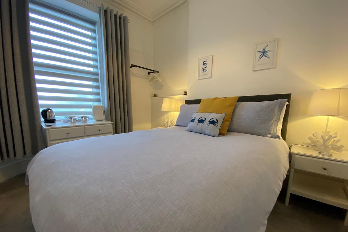 A neatly arranged hotel room with a large bed, a bedside table with a lamp, a nightstand, and a window with blinds.