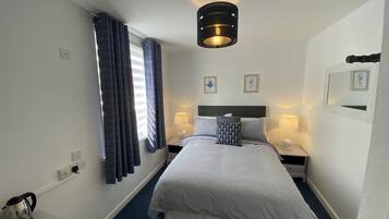 Habitación de hotel con una cama, dos lámparas de noche, una luz de techo, una ventana con cortinas y una mesita con hervidor y tazas.