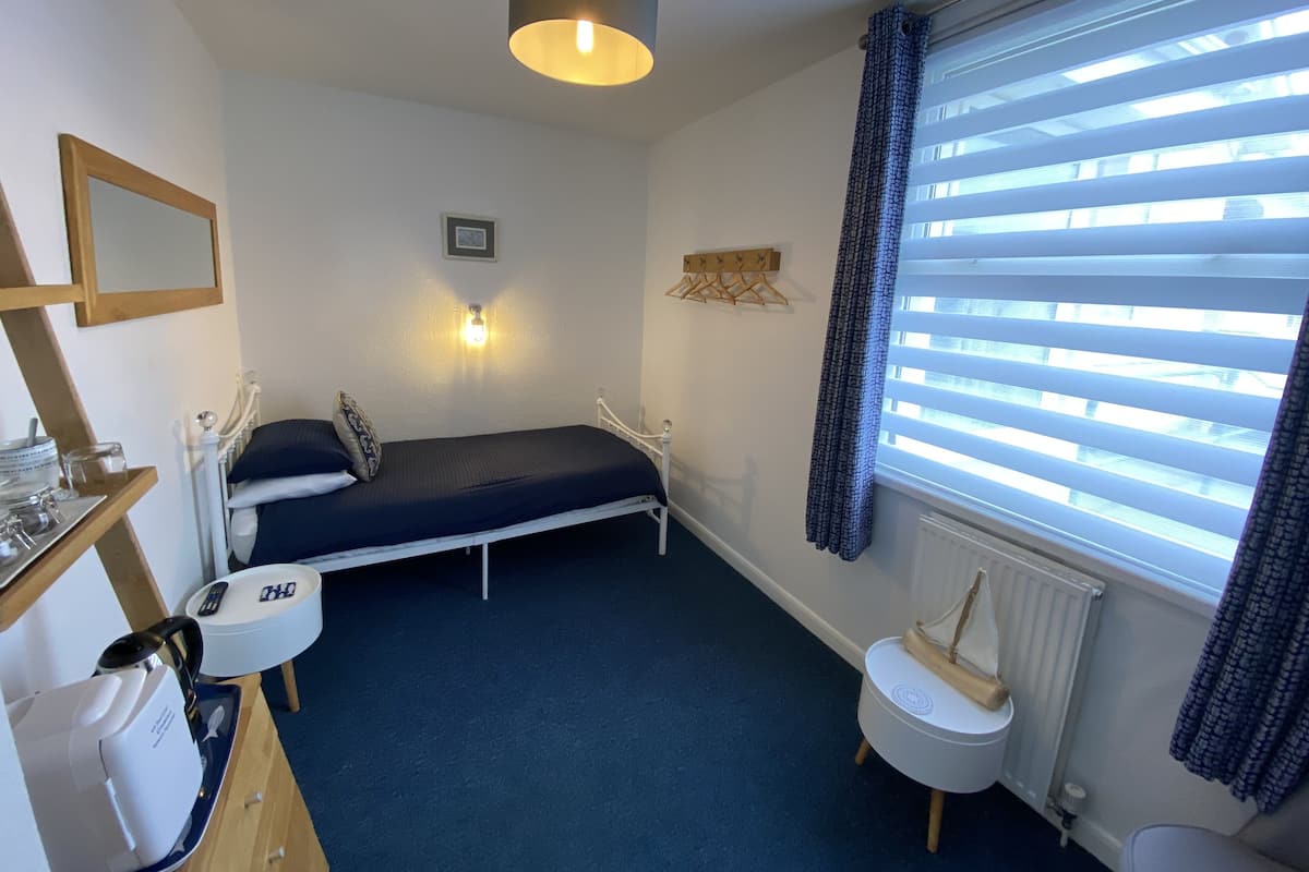 A small, neatly arranged bedroom with a single bed, a bedside table, a small desk, a chair, a radiator, and a window with blinds.