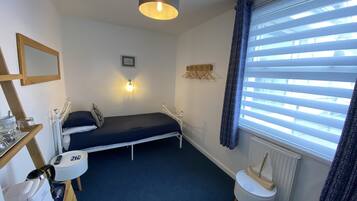A small, neatly arranged bedroom with a single bed, a bedside table, a small desk, a chair, a radiator, and a window with blinds.