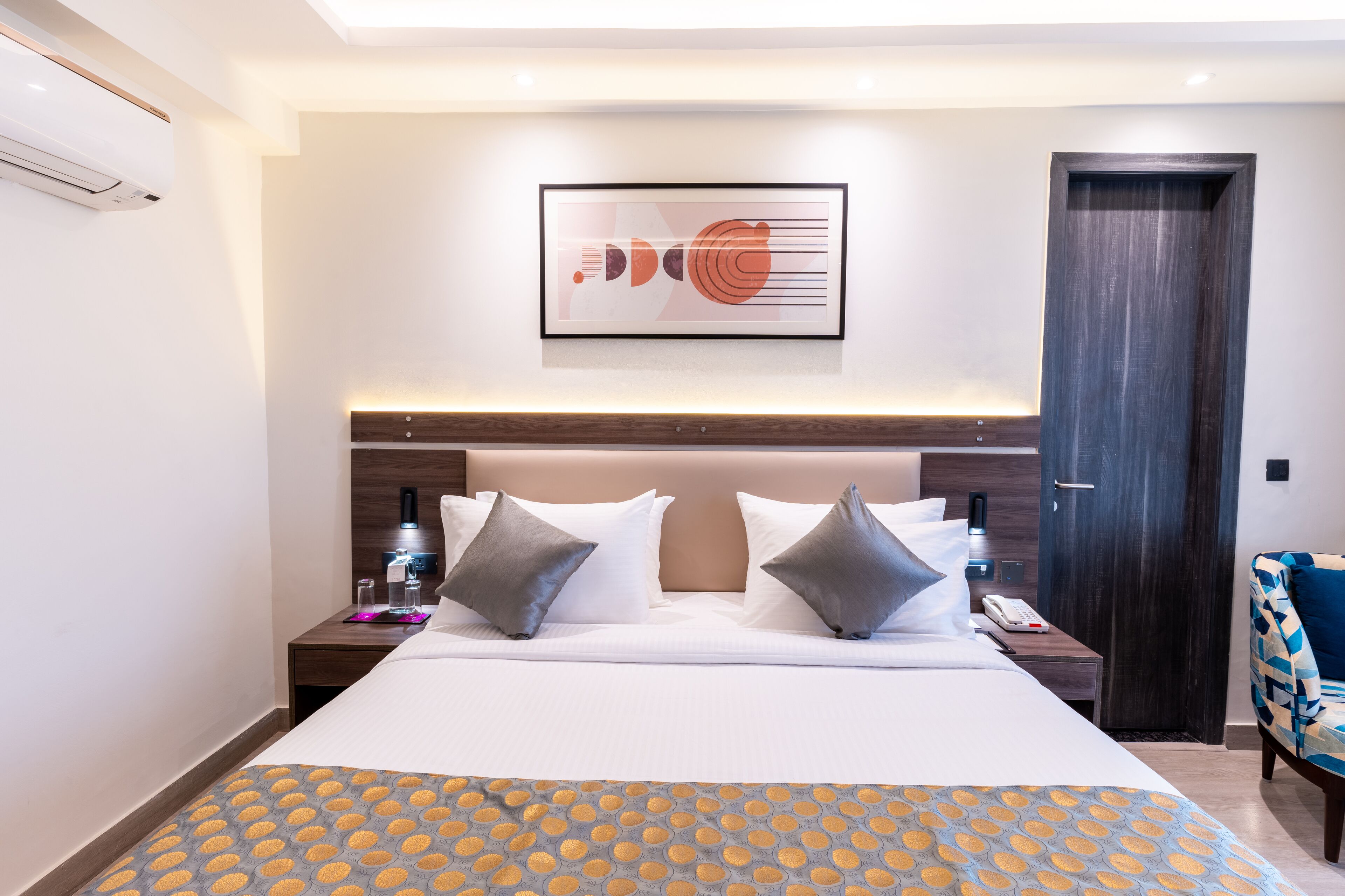 A hotel room with a large bed, a headboard with built-in lighting, a bedside table with a phone and a lamp, and a door to the right.