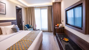 Une chambre d’hôtel avec un grand lit, un bureau agrémenté d’un panier de fruits, une chaise et une grande fenêtre avec des rideaux.