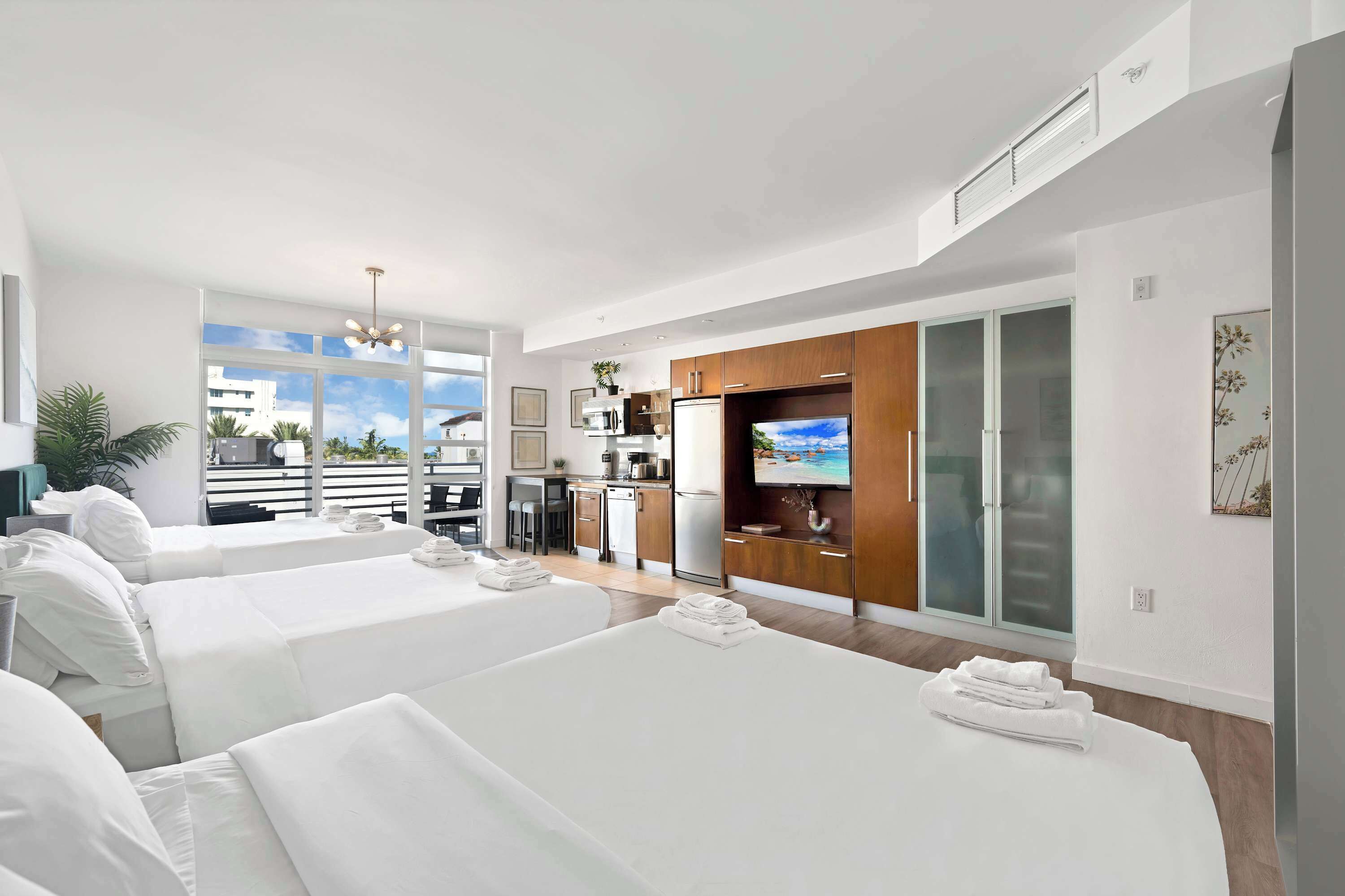 Une chambre d’hôtel moderne avec deux lits, une kitchenette, une télévision et un balcon offrant une vue sur la ville.