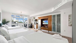 Habitación de hotel moderna con dos camas, kitchenette, televisión y balcón con vistas a la ciudad.