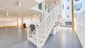 Un aparcamiento de varios niveles con una escalera blanca, un letrero de salida de emergencia rojo y un edificio con múltiples ventanas al fondo.