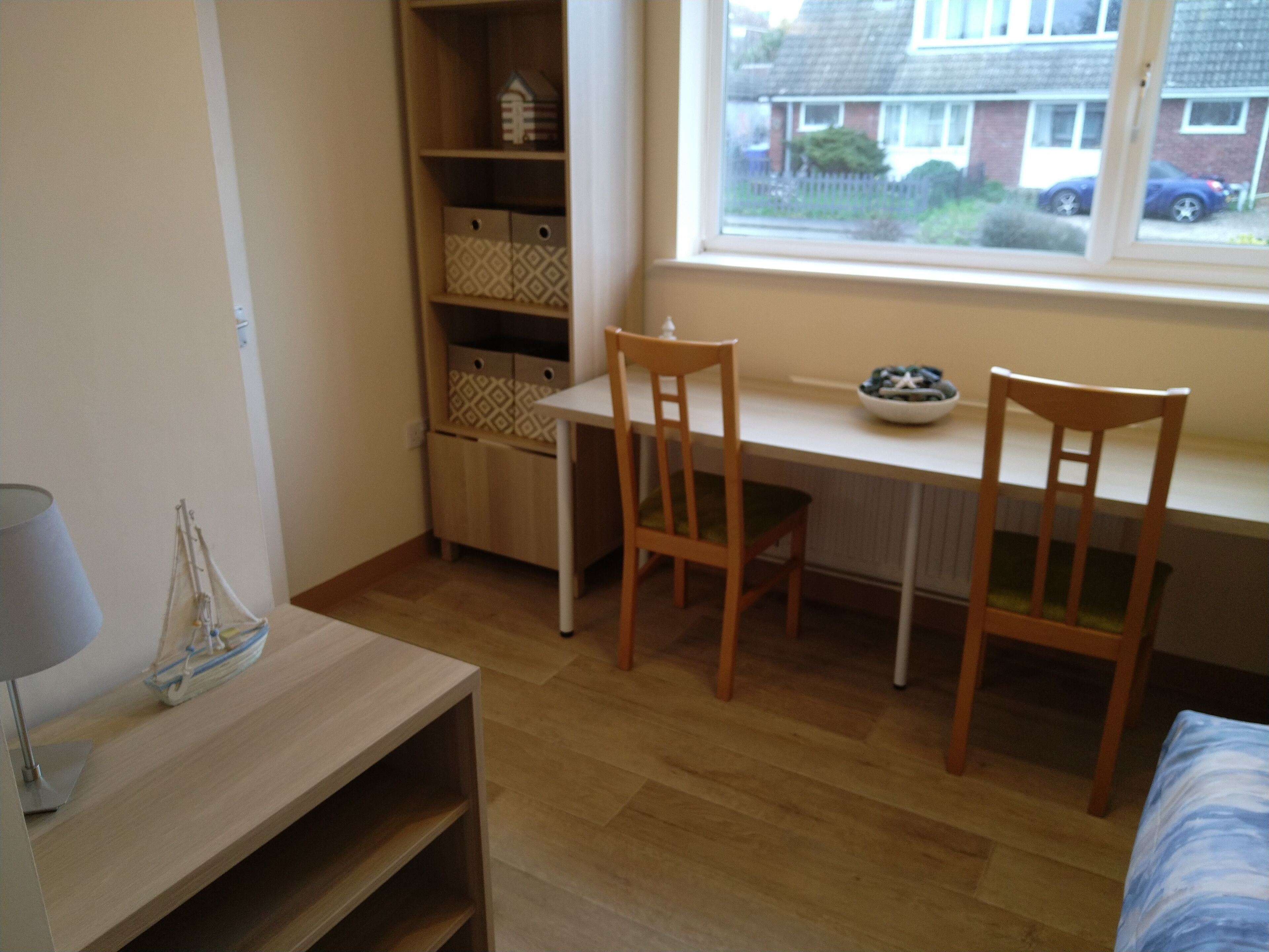 A room with a wooden desk, two chairs, a lamp, and a window with a view of houses.