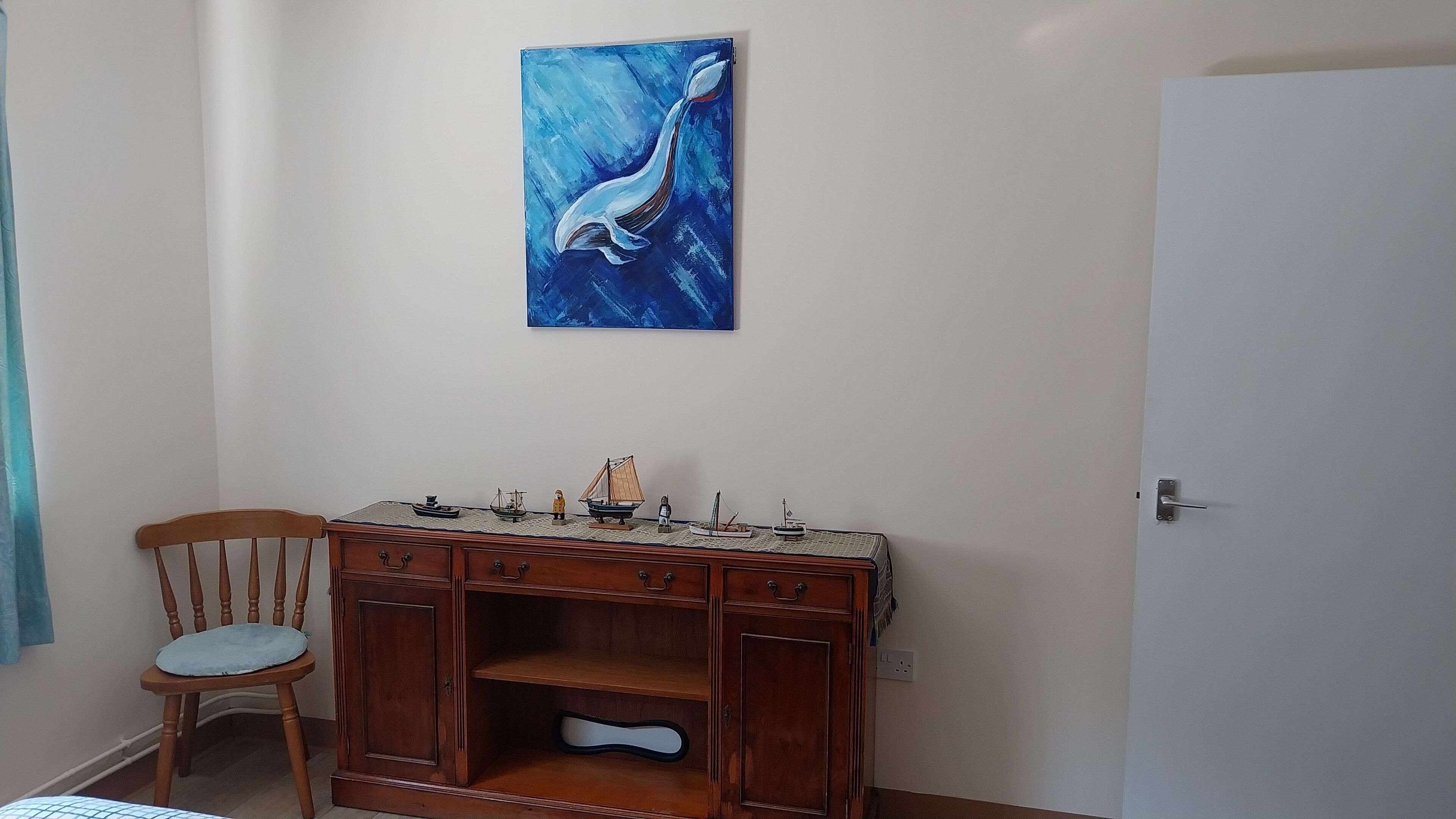A room with a wooden cabinet displaying model ships, a chair, and a painting of a whale on the wall.