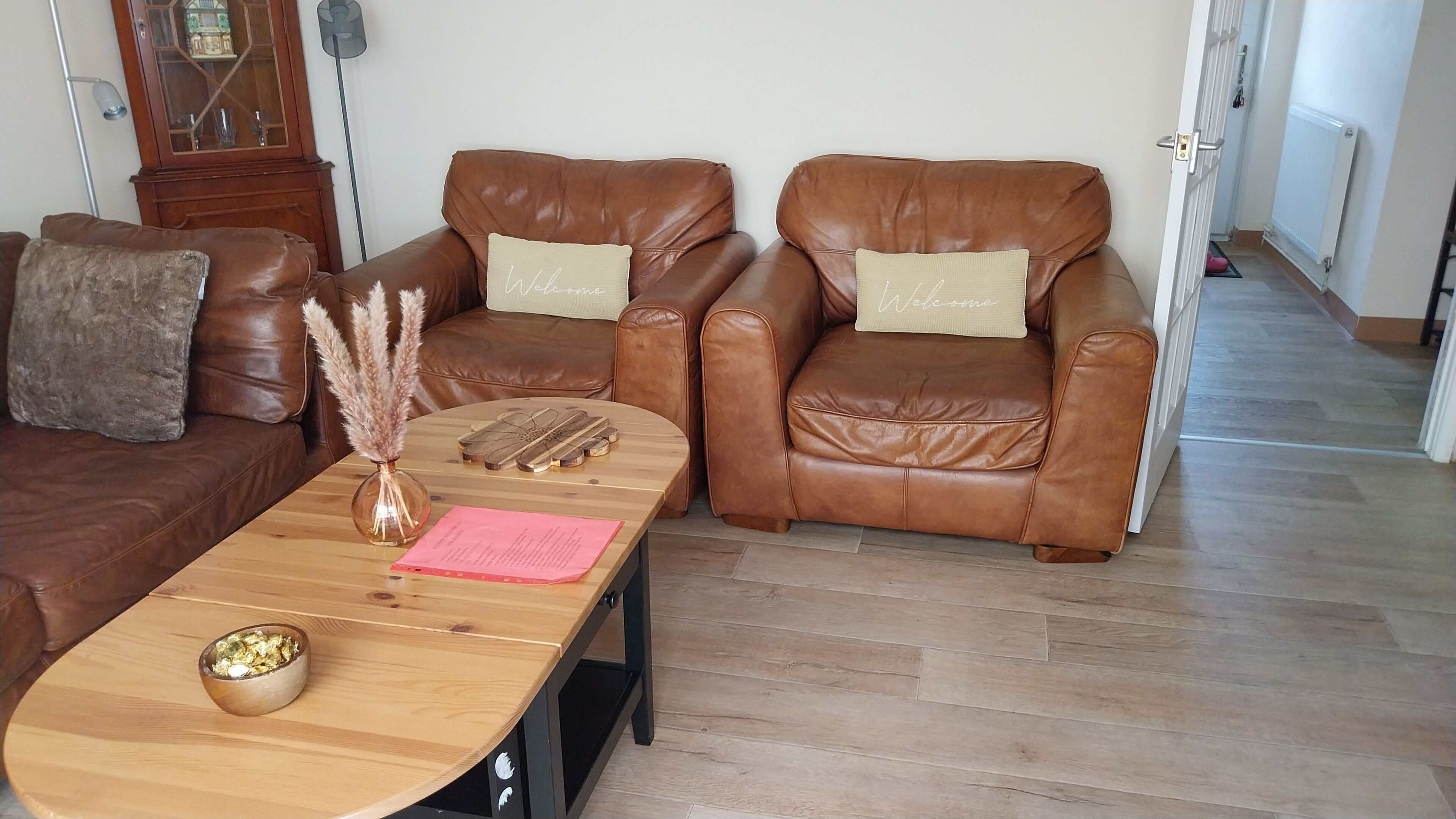 A living room with a brown leather sofa, two brown leather armchairs, a wooden coffee table, and a vase with pampas grass.