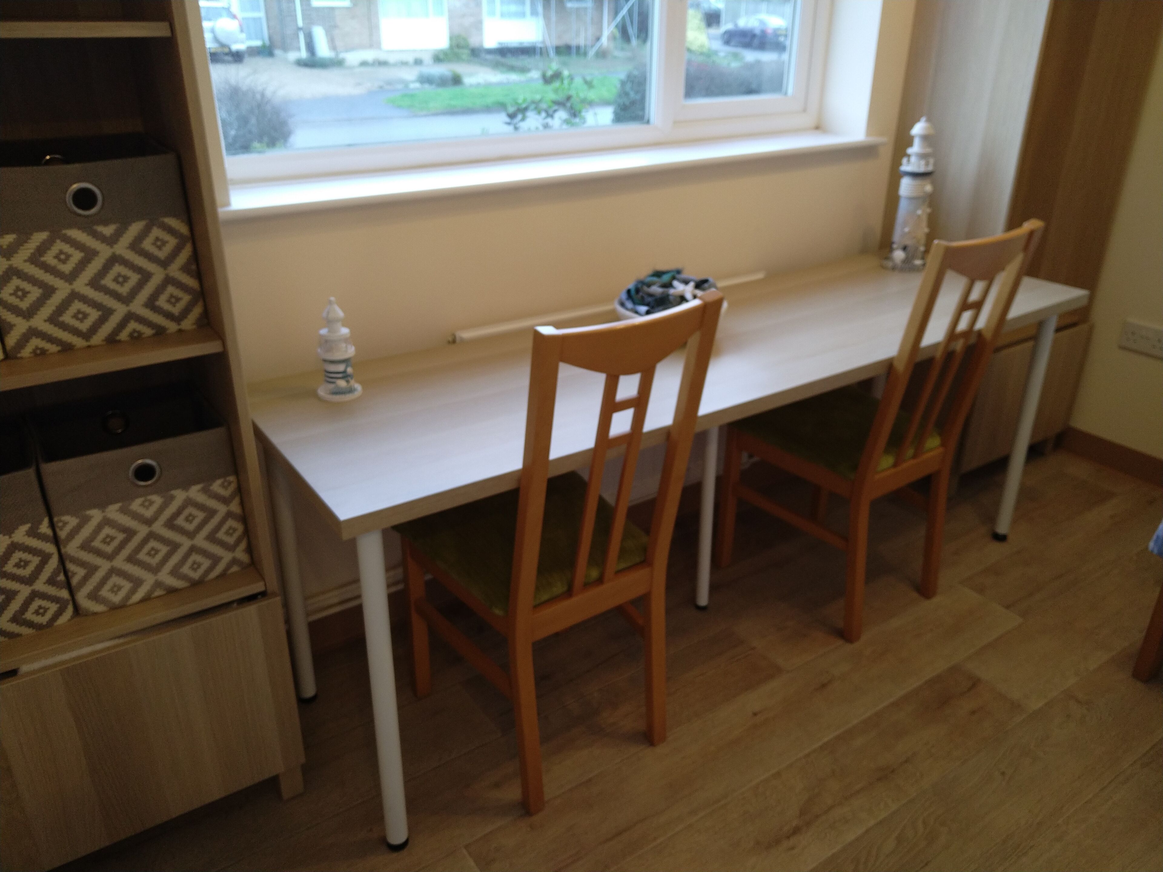 A small dining area with a white table and two wooden chairs, a window with a view of the outside, and a patterned storage unit to the left.