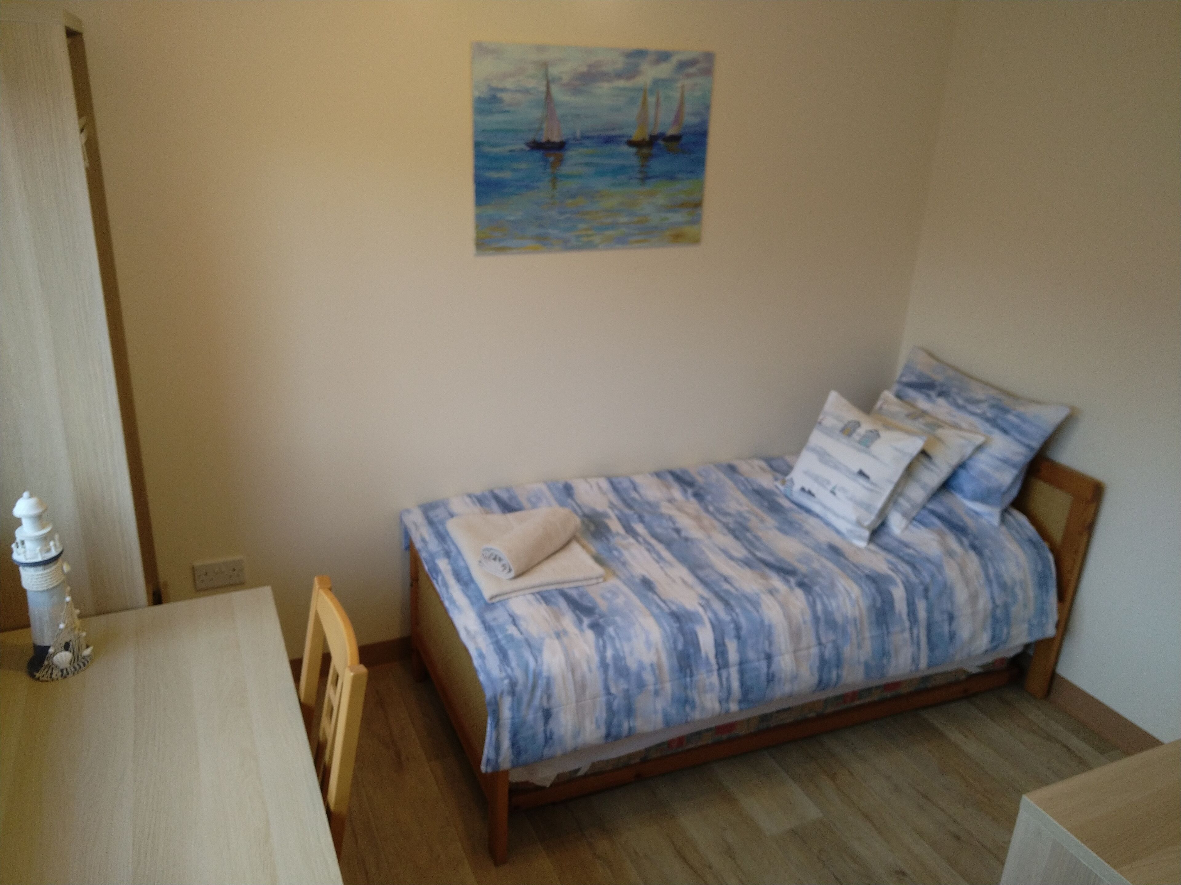 A small, neatly arranged bedroom with a single bed, a wooden nightstand, a small table, and a wall-mounted painting of sailboats.