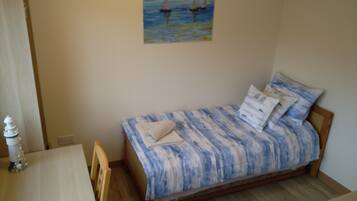 A small, neatly arranged bedroom with a single bed, a wooden nightstand, a small table, and a wall-mounted painting of sailboats.