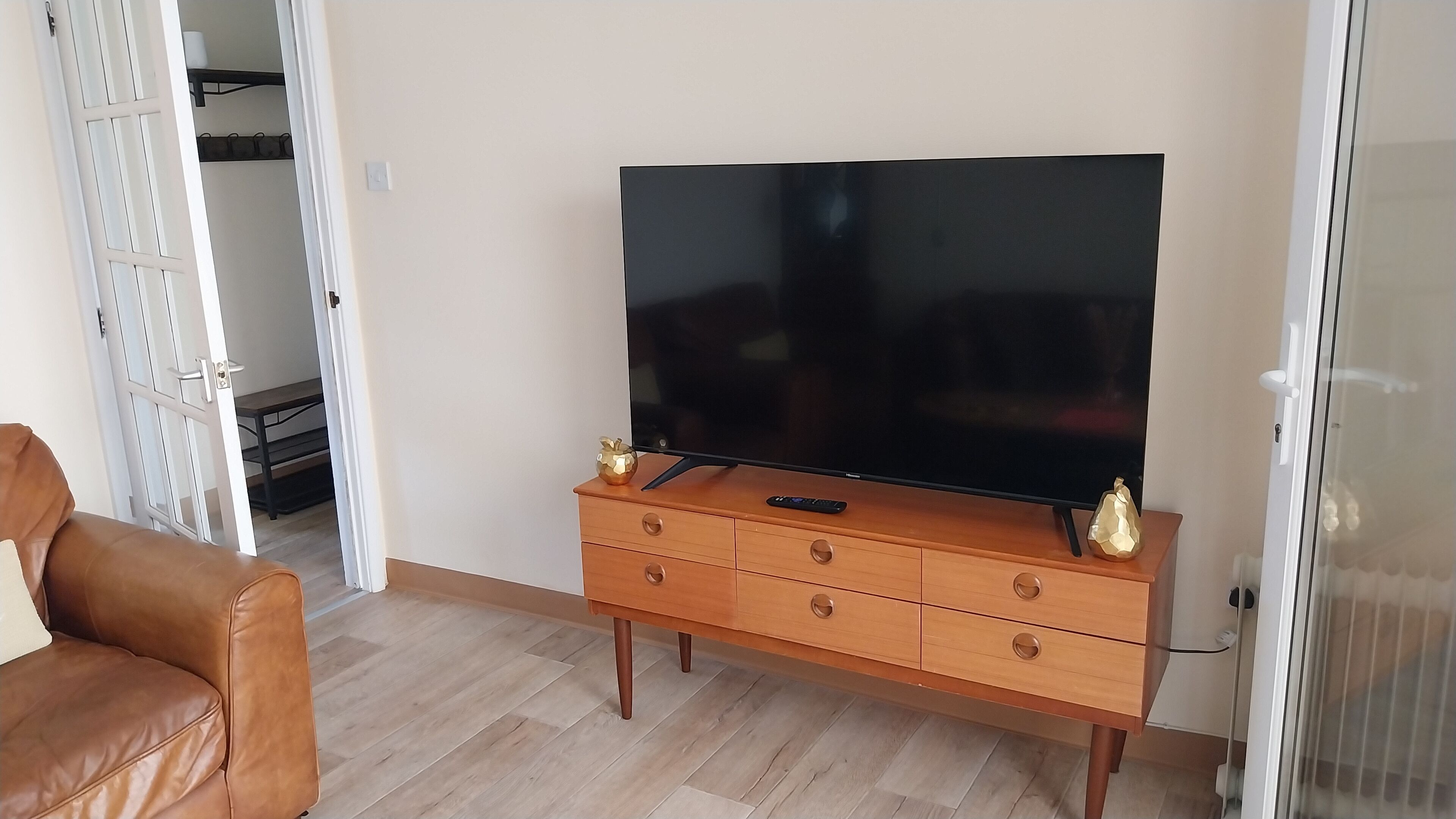 Olohuoneessa on litteänäyttöinen televisio, joka on sijoitettu puiseen kaappiin, ruskea nahkasohva ja lasiovi, joka johtaa toiseen huoneeseen.