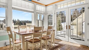 Un comedor con mesa y sillas de madera, ventanas amplias con persianas y vistas a un paisaje nevado.