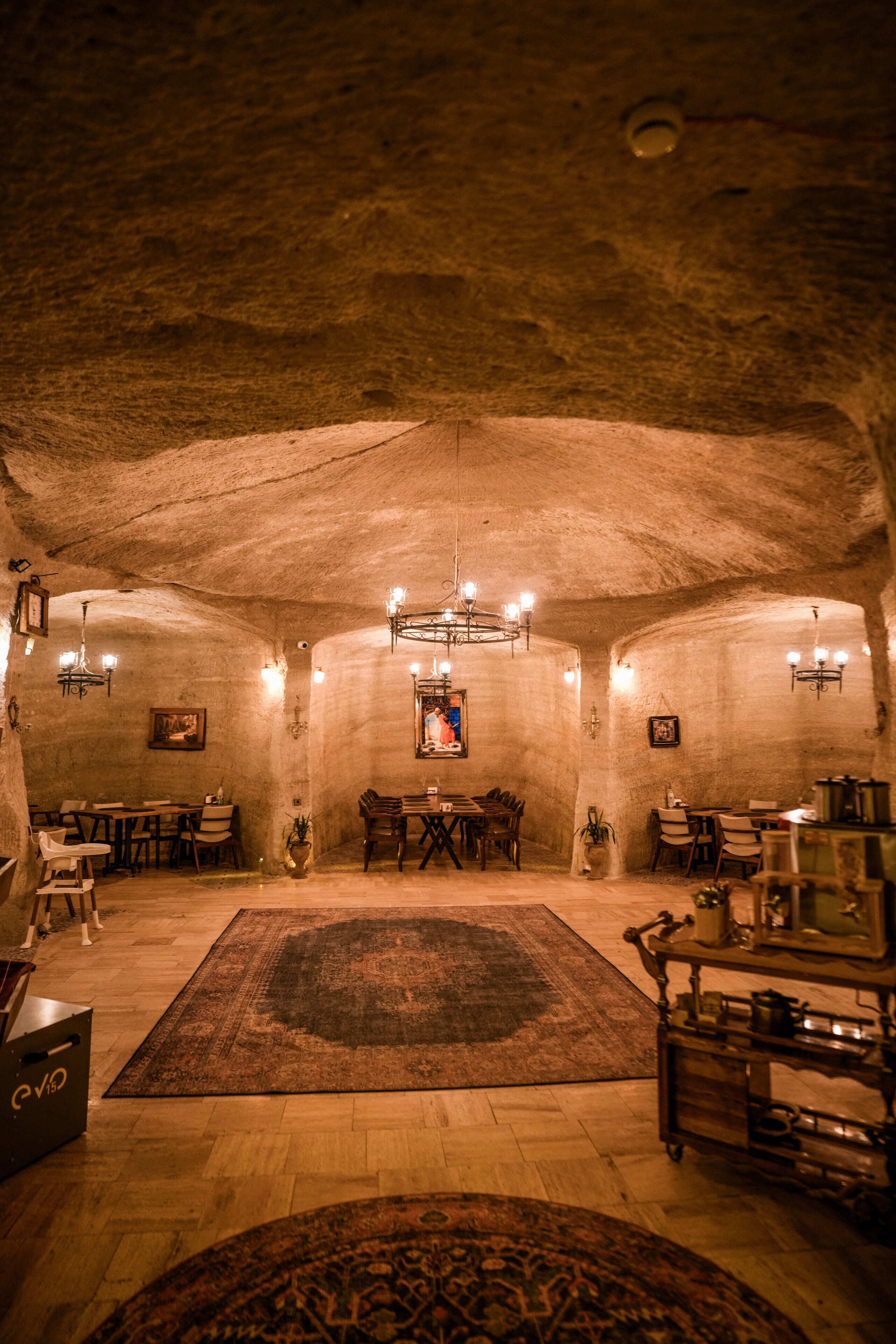 A spacious underground dining area with wooden tables, chairs, and a central chandelier.