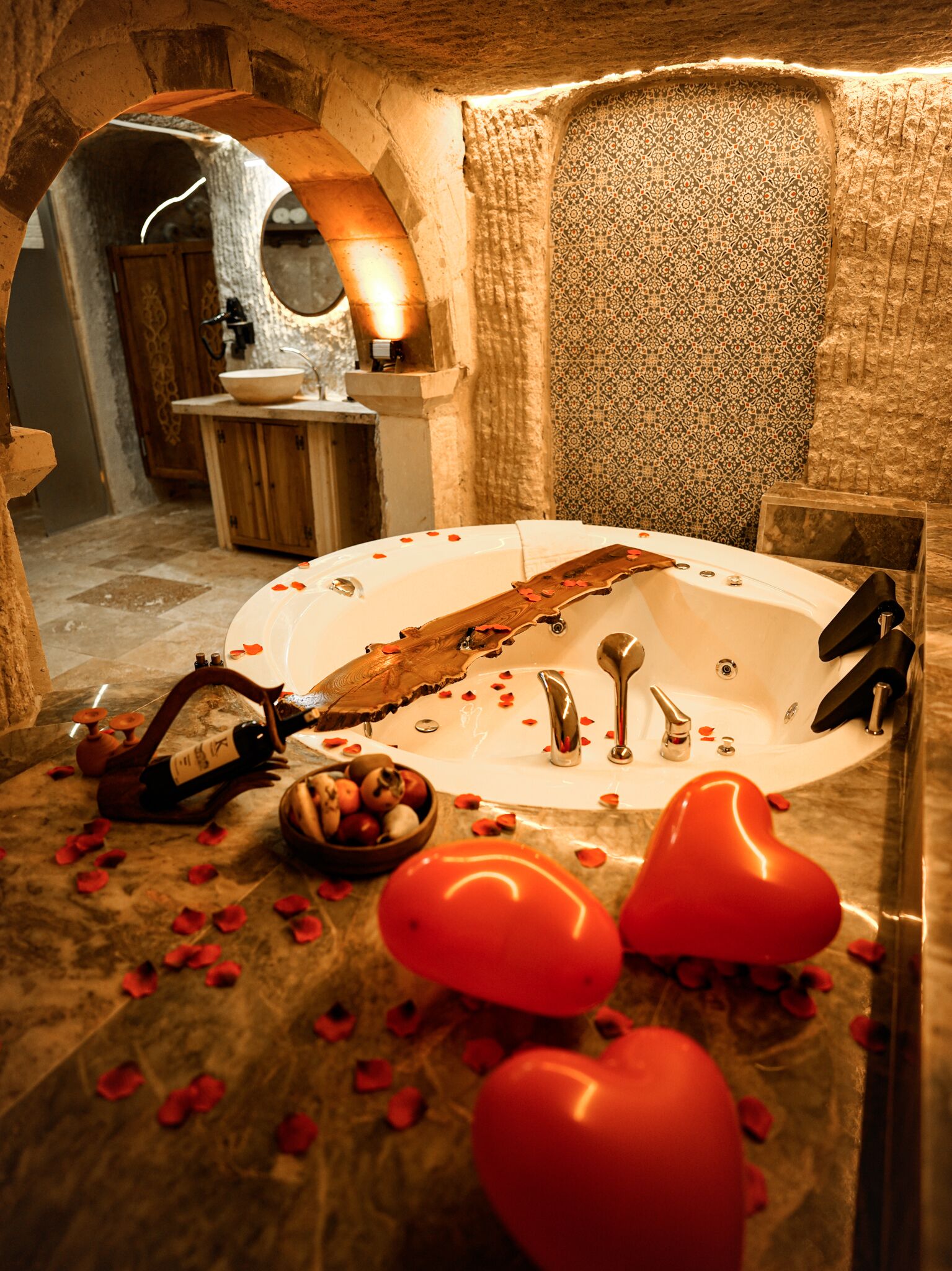 A bathroom with a bathtub, a wooden wine bottle, a bowl of fruit, and heart-shaped decorations.