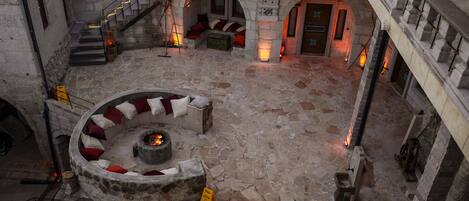 Un patio con suelo de piedra, una zona de estar circular con cojines y una chimenea. Se observan paredes de piedra y una escalera que conduce a un nivel superior.