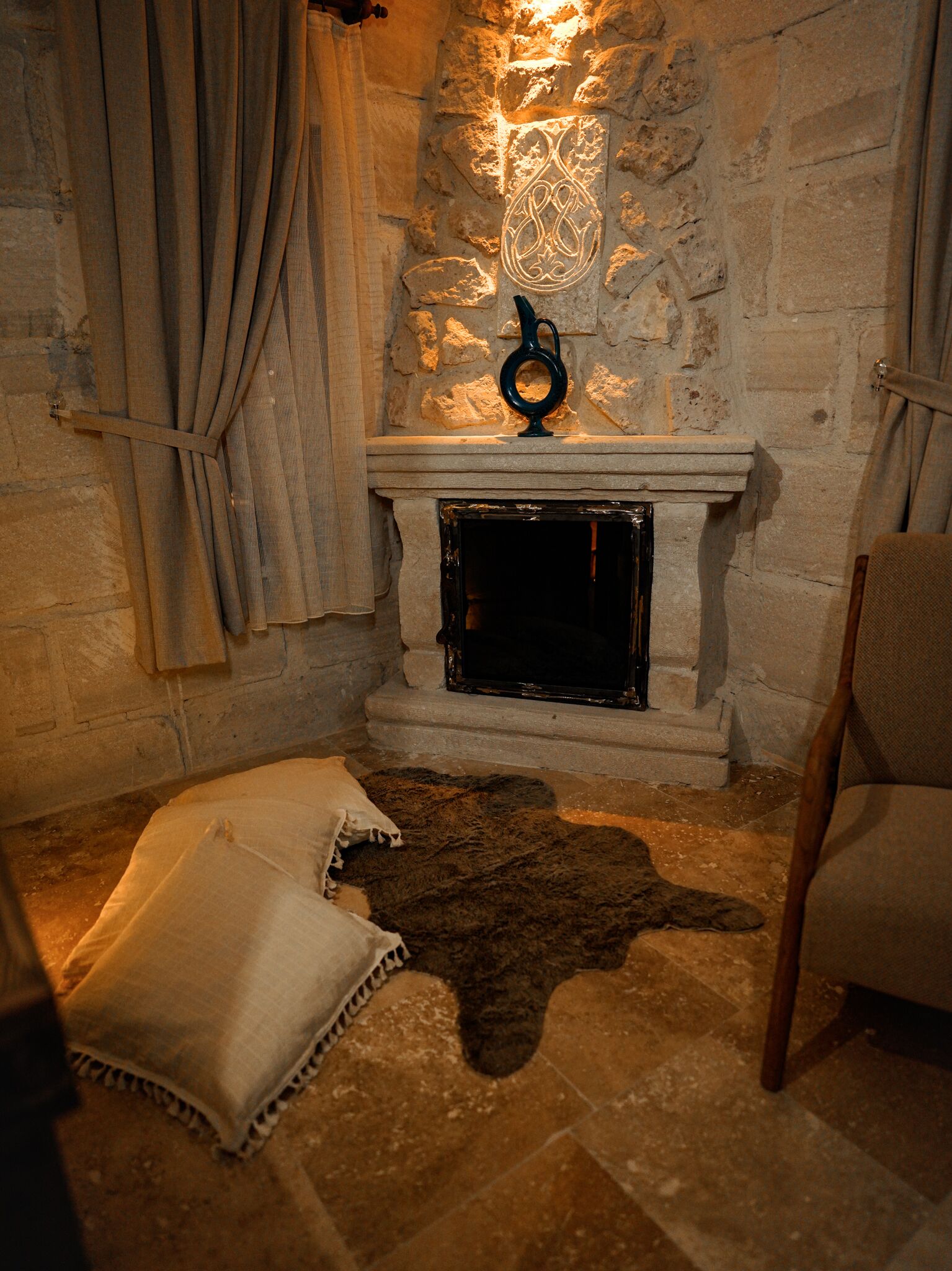 Una chimenea de piedra con un adorno metálico encima, un tapete de piel en el suelo y una silla a la derecha.