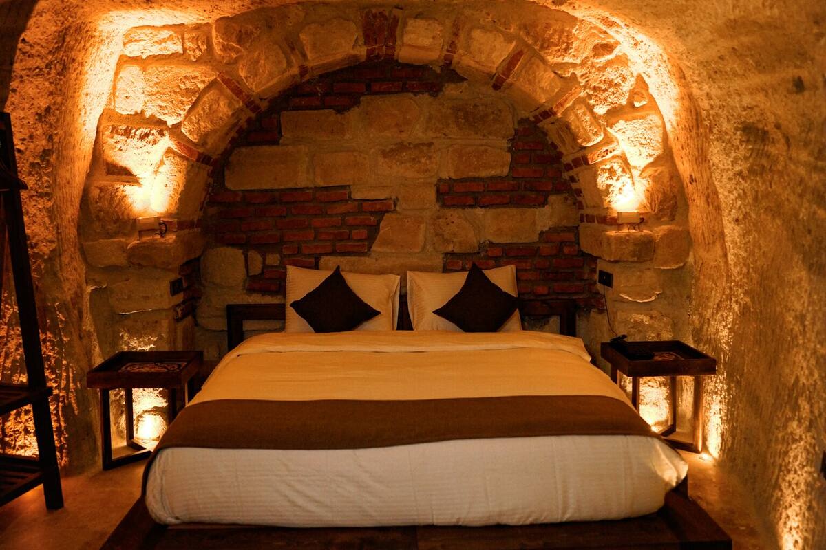 Un llit en una habitació amb parets de pedra i un sostre amb arc de rajola, dues taules de nit amb fanalets i un terra de fusta.