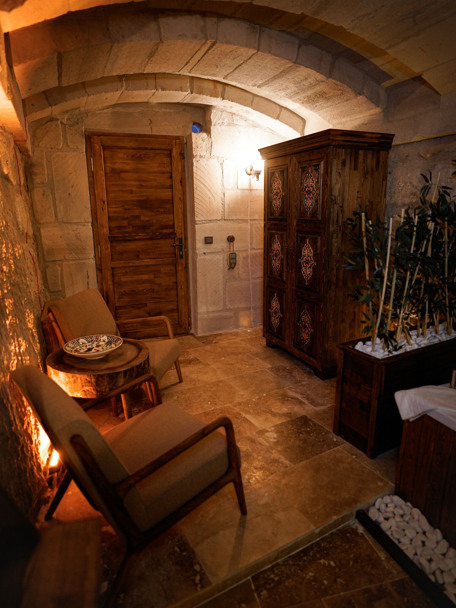 A stone-walled room with a wooden door, a cabinet with intricate designs, a chair, a small table with a decorative plate, and a potted plant.