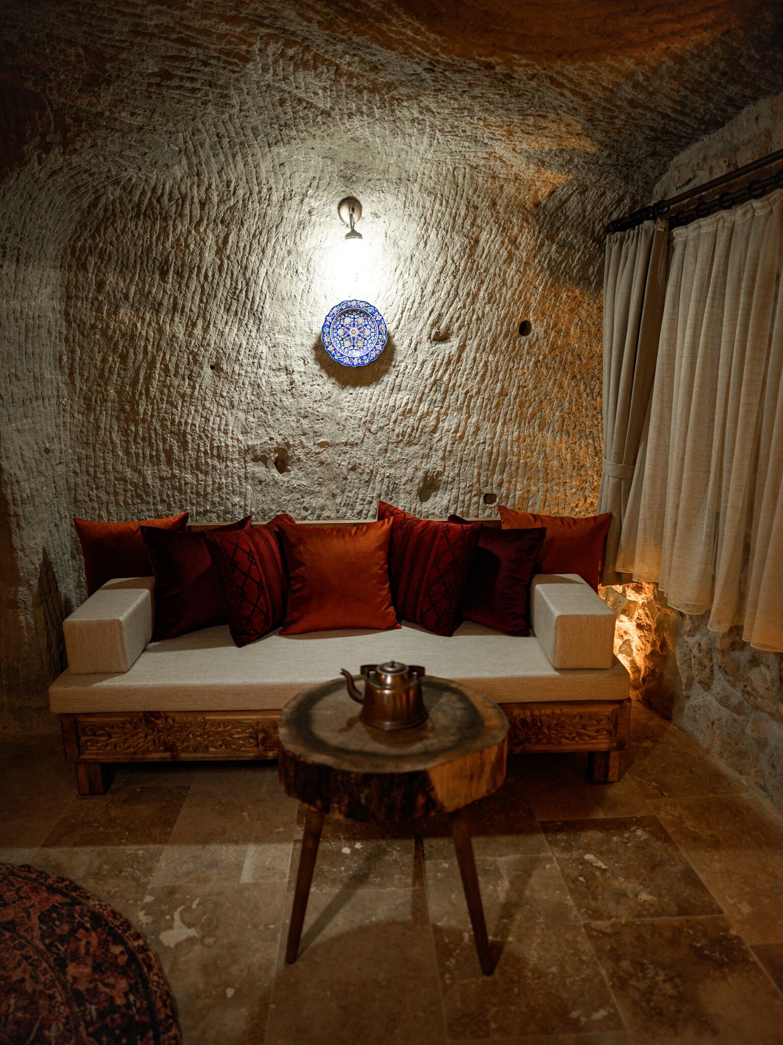 A cozy room with a stone wall, a sofa with cushions, a small wooden table with a teapot, and a decorative plate on the wall.