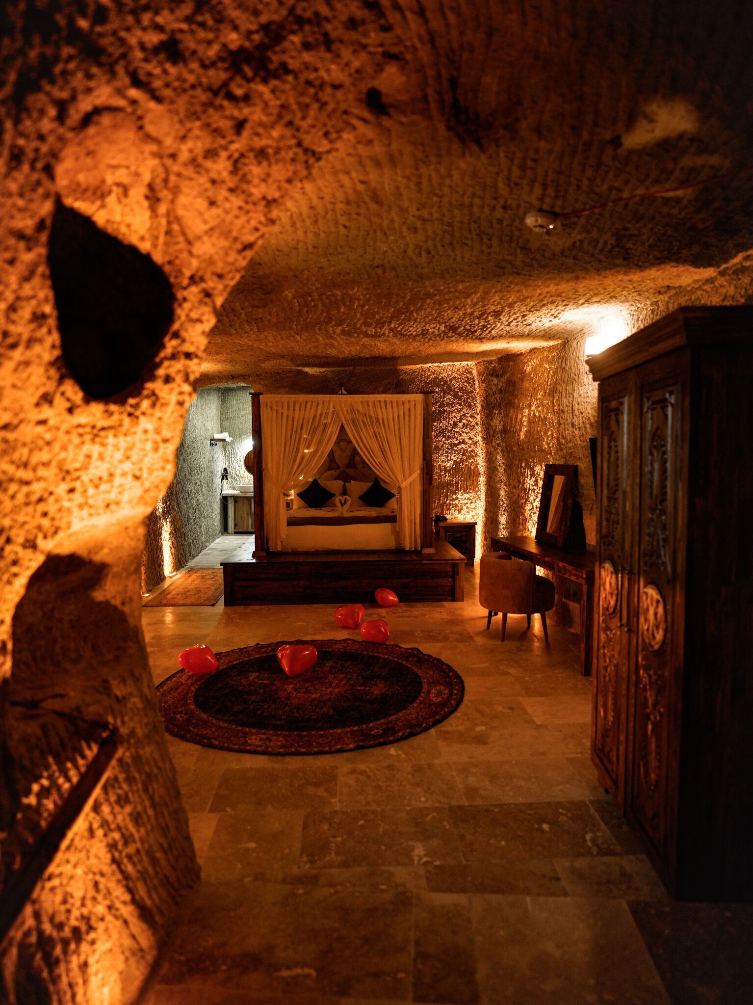 A cozy interior with a stone wall, a bed with draped curtains, a wooden cabinet, and a round rug with heart-shaped objects.