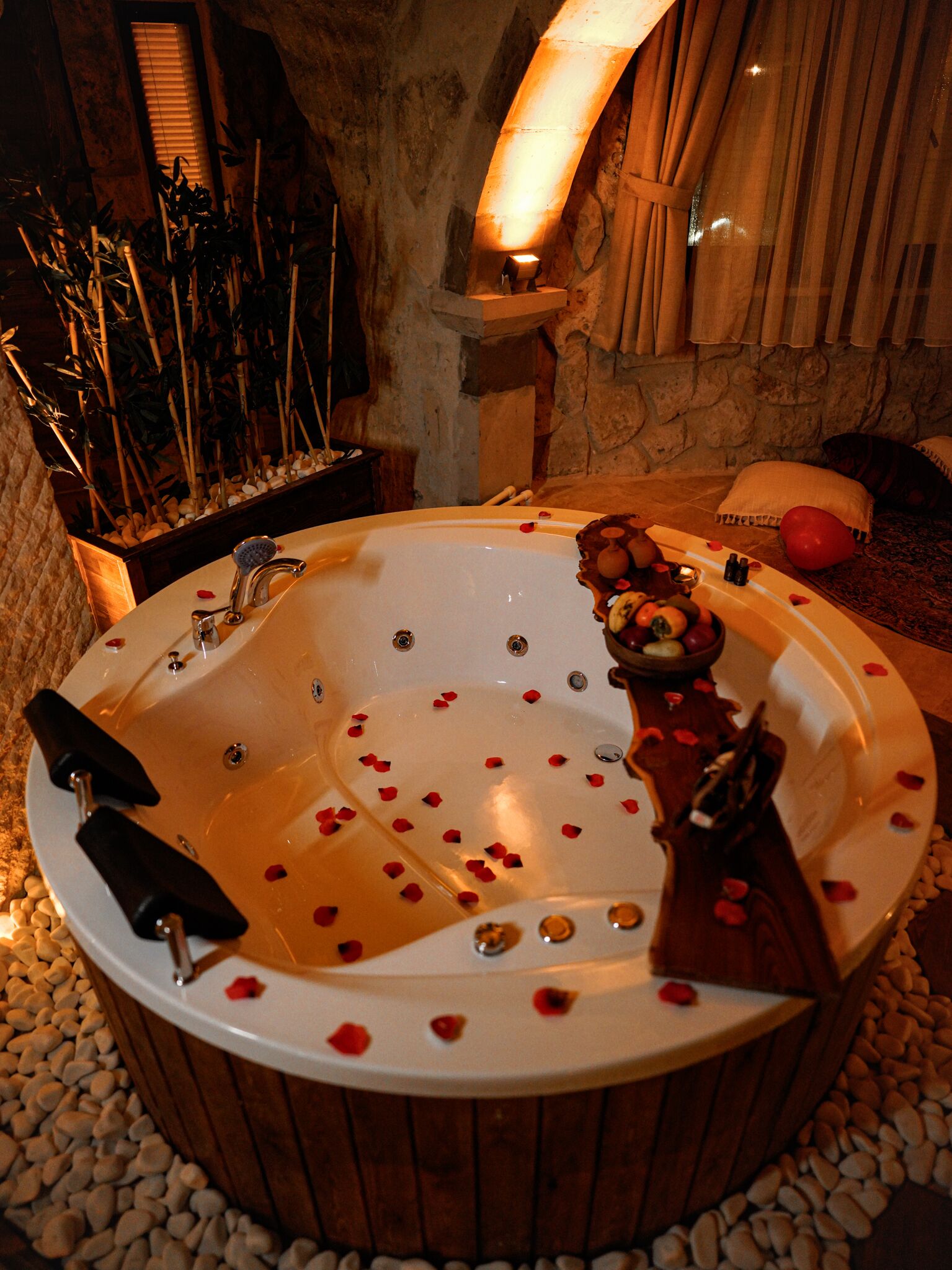 Un jacuzzi con pétalos de rosa, rodeado de piedrecitas, en un cuarto de paredes de piedra con un banco de madera y un estante pequeño con plantas.