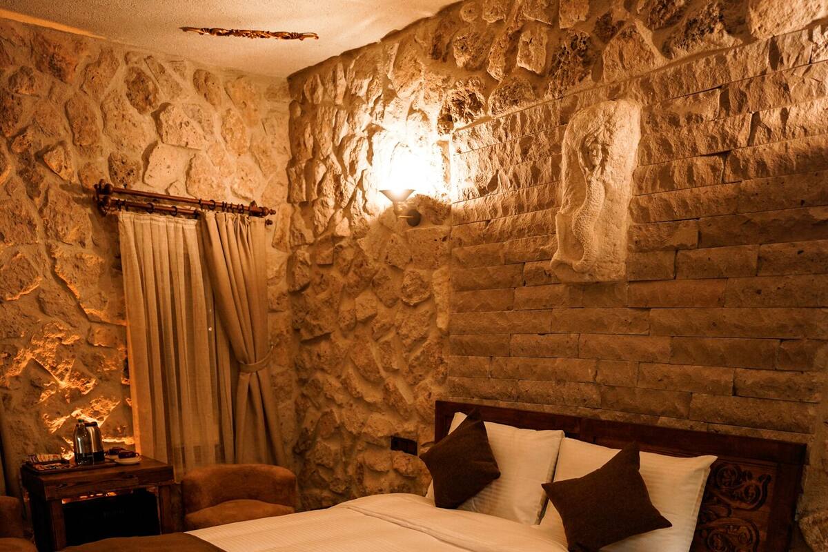 Un dormitori amb paret de pedra, llit de fusta amb capçalera, una tauleta de nit, una làmpada i una finestra amb cortines.