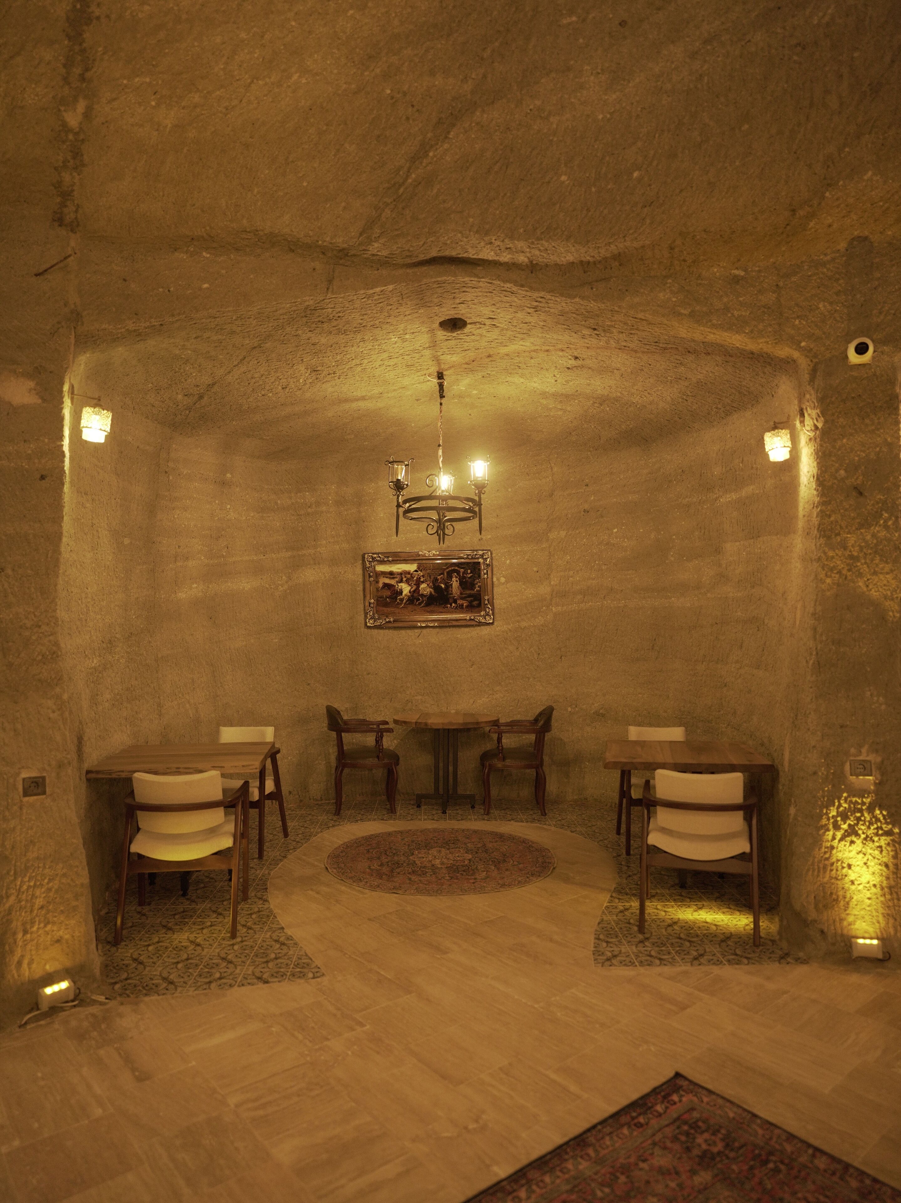 A cozy, dimly lit room with a rustic stone wall, a wooden table with chairs, a chandelier, and a framed picture on the wall.