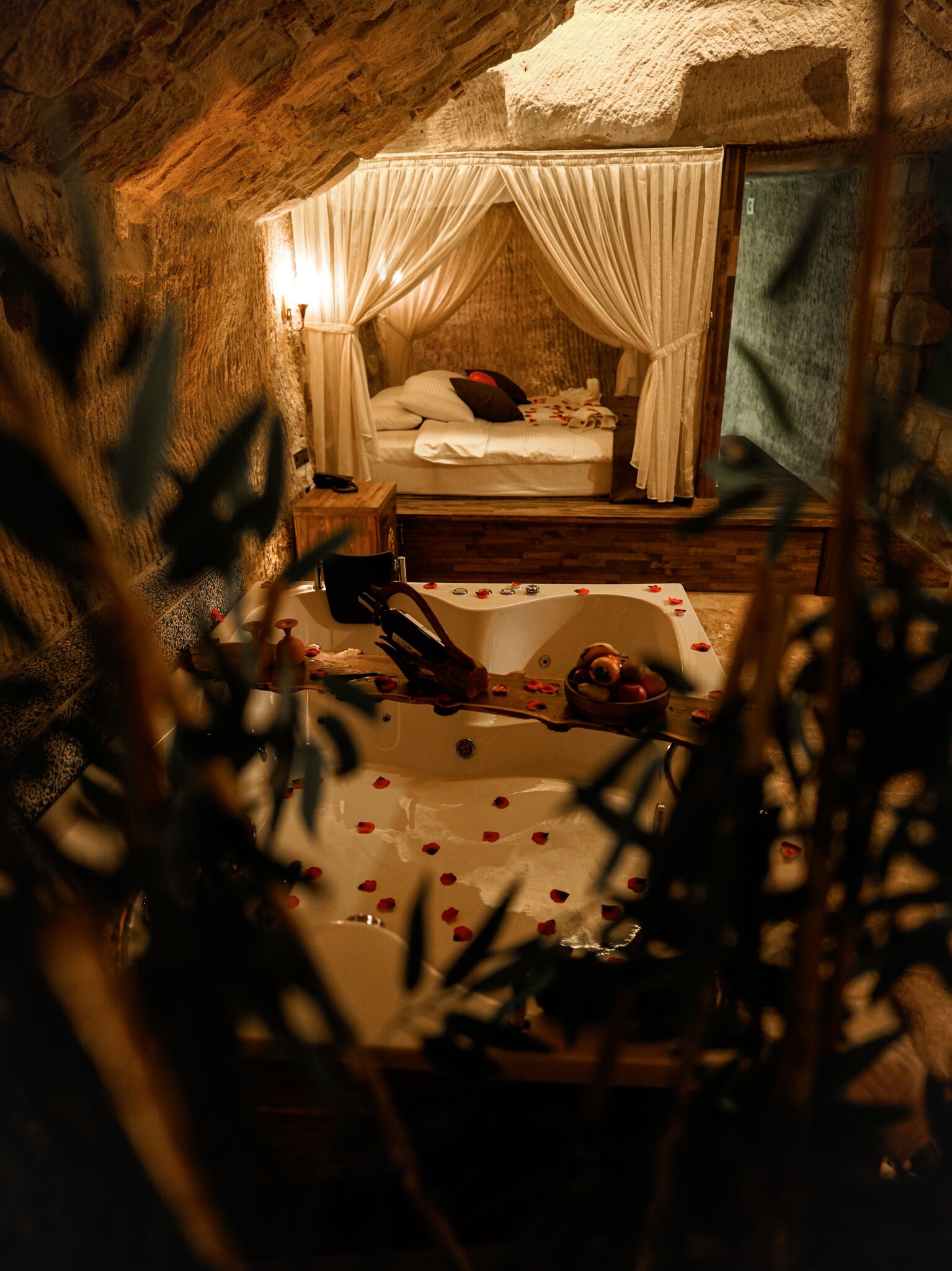 A rustic stone room with a bed, a bathtub with rose petals, and a wooden bench.