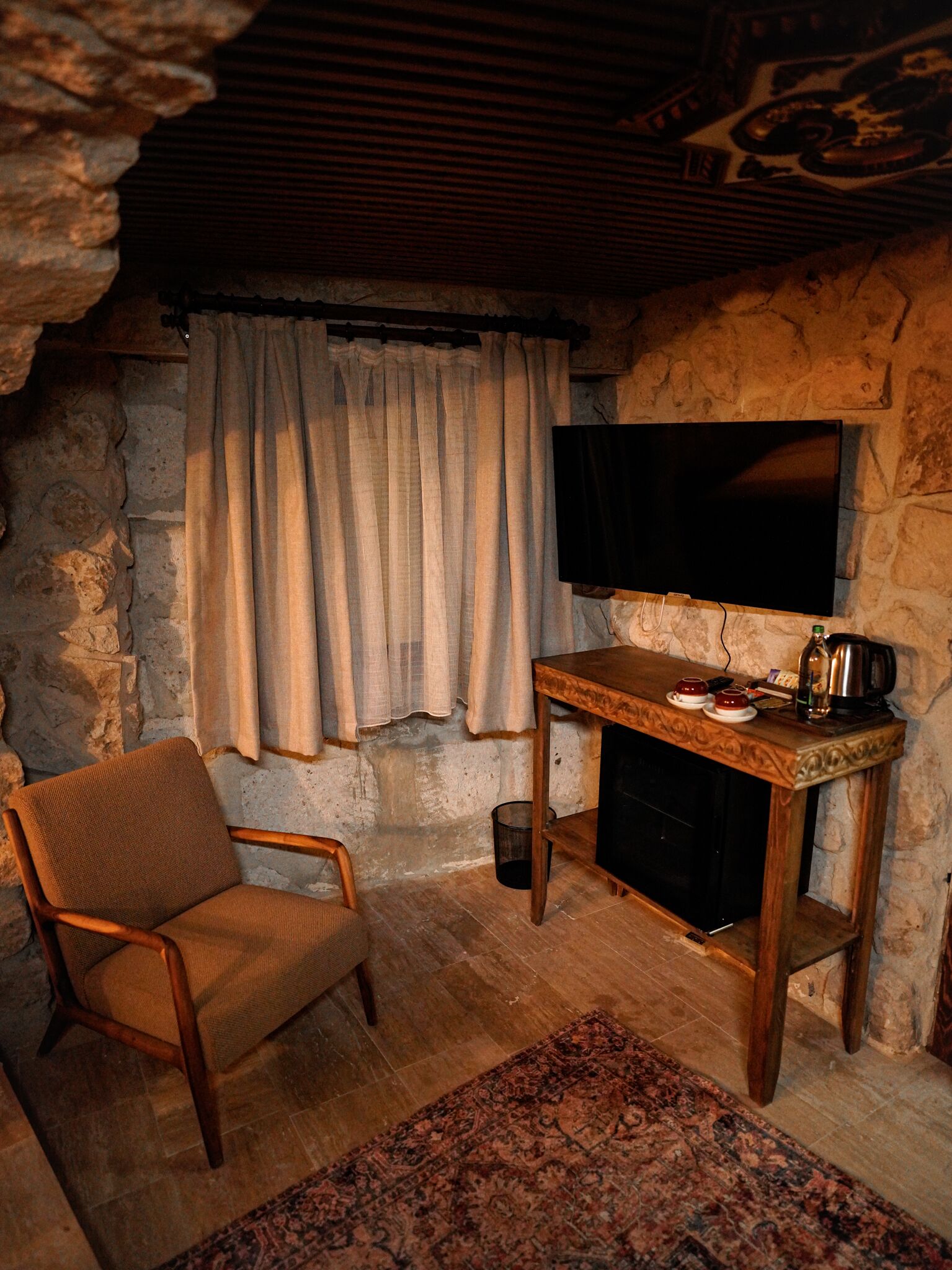 A cozy room with a stone wall, a wooden chair, a flat-screen TV on a wooden stand, and a small table with a kettle and cups.