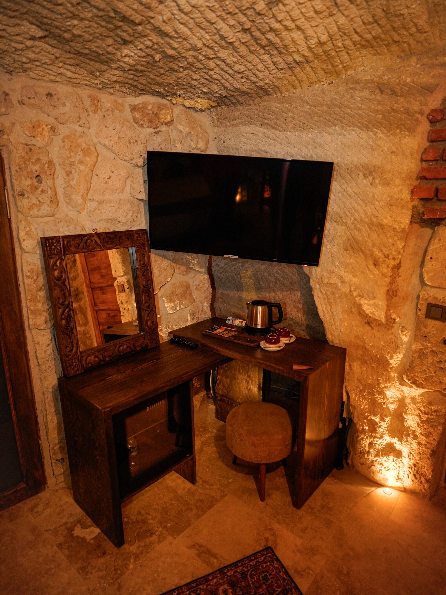 A stone-walled room with a wooden desk, a television mounted on the wall, and a small table with a kettle and cups.