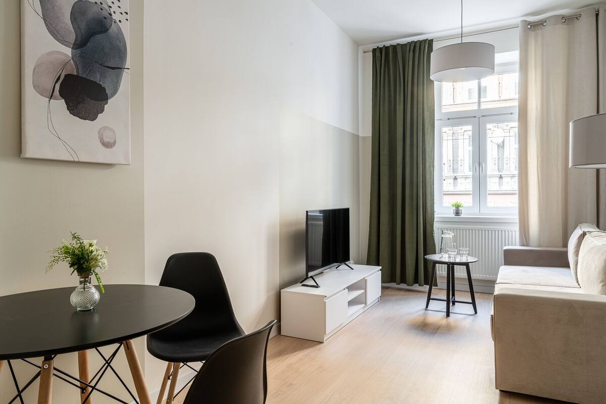 A modern living room with a flat-screen TV, a dining table with chairs, a sofa, and a large window with curtains.
