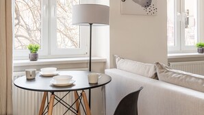 Ein modernes Wohnzimmer mit einem weißen Sofa, einem runden Couchtisch und einem schwarzen Sessel. Eine Stehlampe, ein Gemälde an der Wand und ein Fenster mit Blick auf kahle Bäume sind ebenfalls zu sehen.