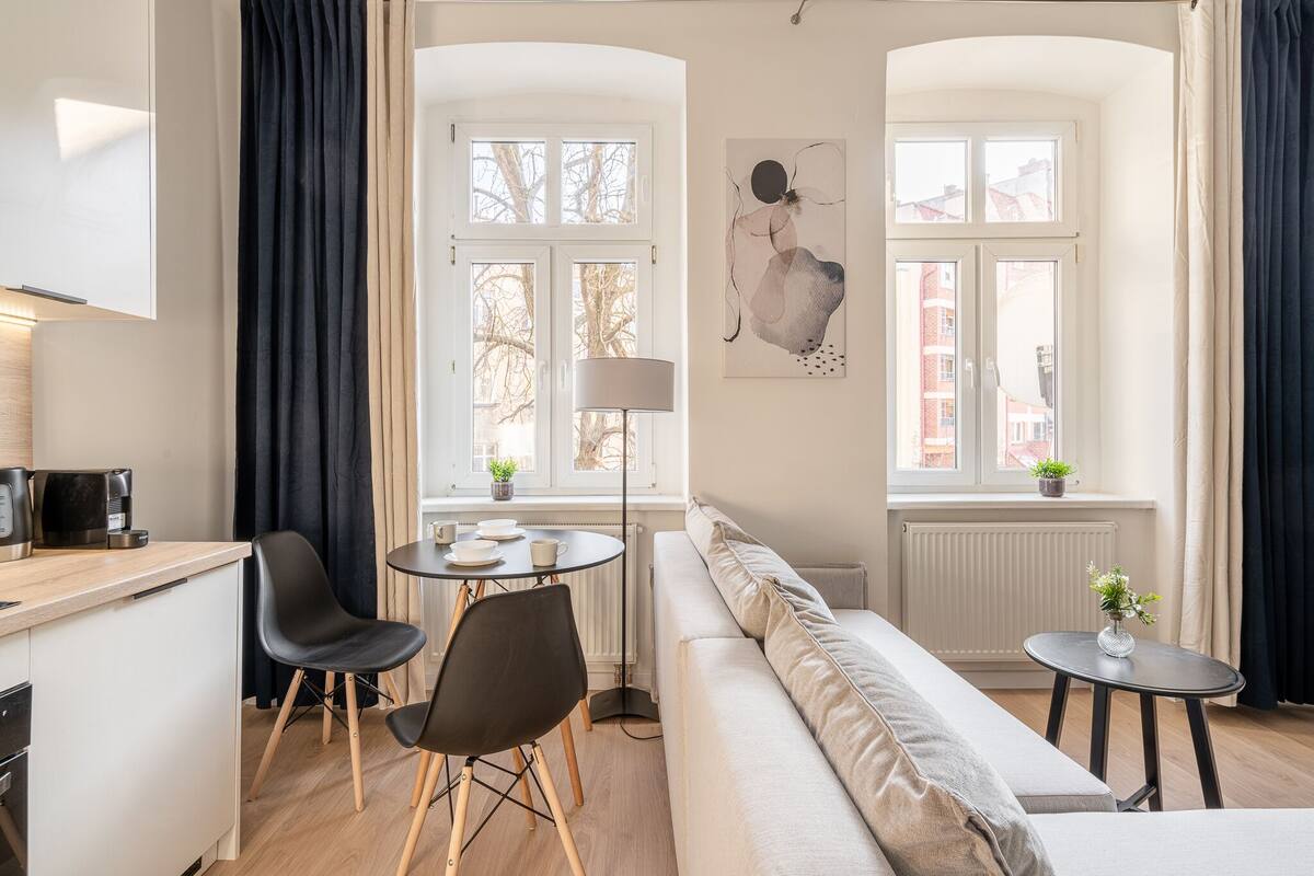 A modern living room with a sofa, a dining table with chairs, a coffee machine, and a window with curtains.