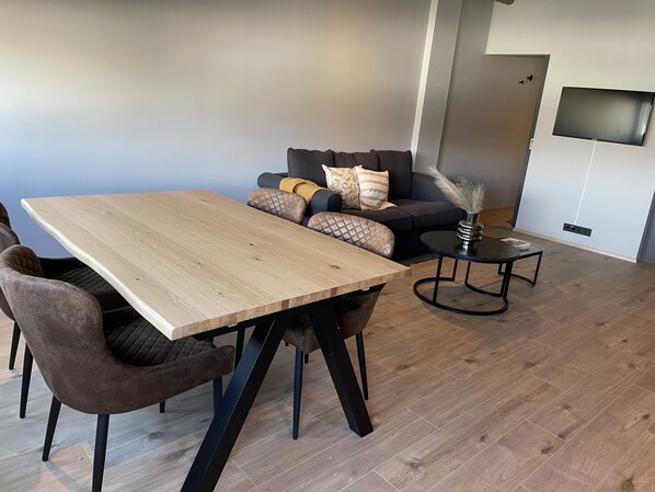 A modern living room with a wooden dining table, cushioned seating, a small side table, and a flat-screen TV mounted on the wall.