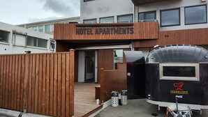 Entrada de um hotel com fachada de madeira, uma placa com a inscrição "HOTEL APARTMENTS" e uma trailer preta com o logotipo da Coca-Cola.